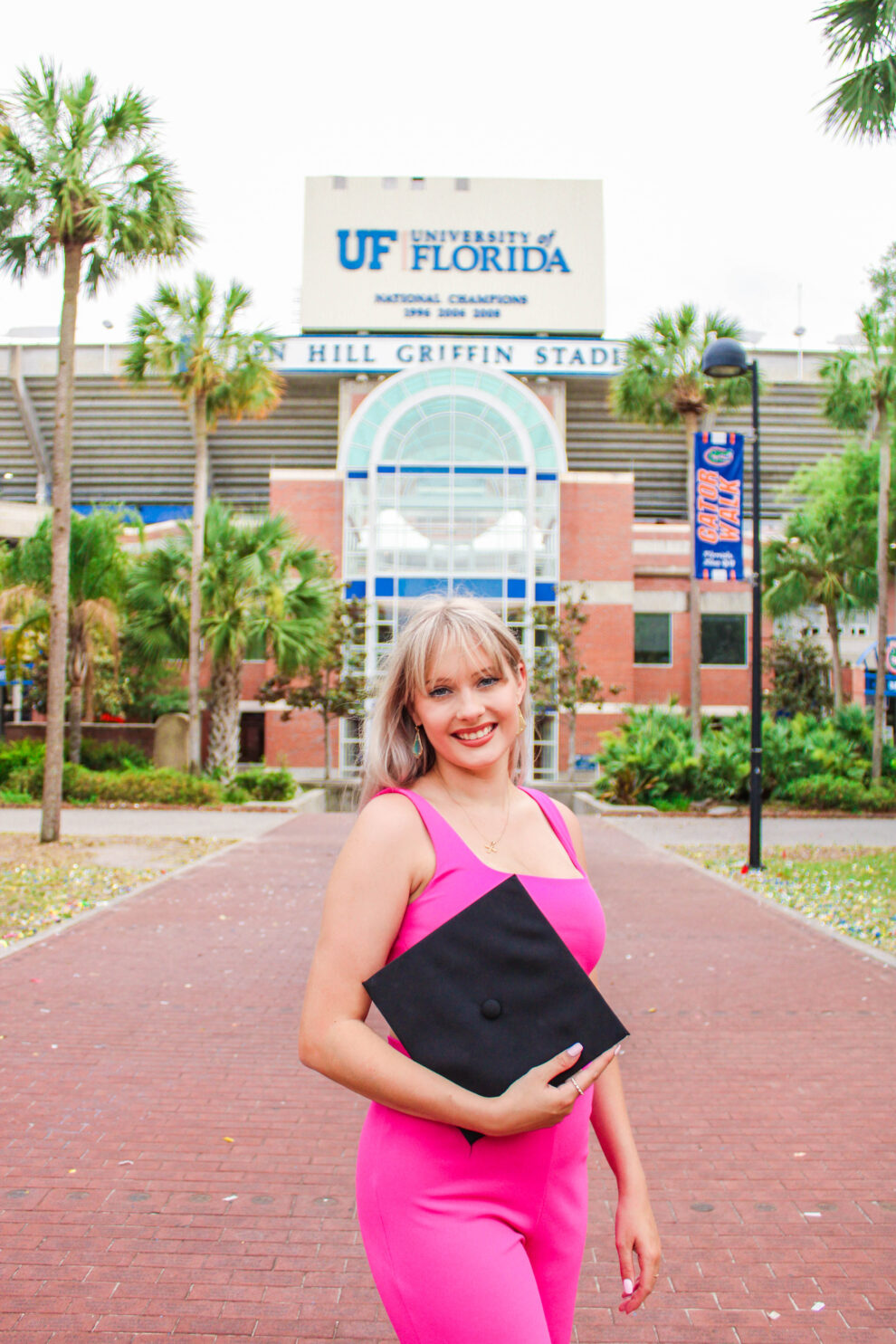 A woman in a pink dress, holding a square object, smiles as she stands sideways to the camera.