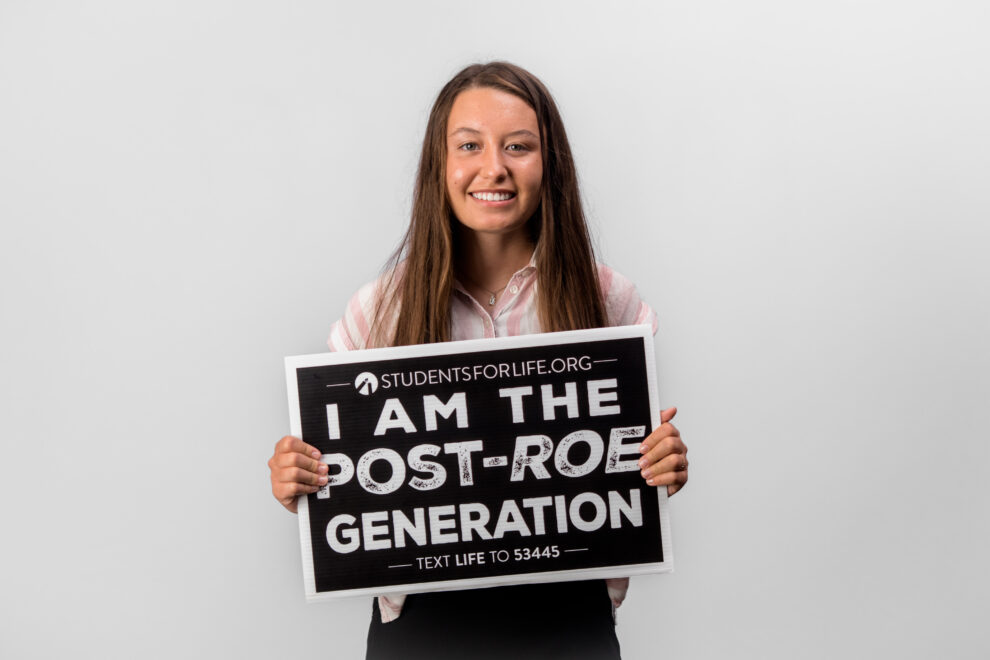 A woman with long brown hair smiles at the camera. She is holding a sign that reads, "I AM THE PRO-LIFE GENERATION. TEXT LIFE TO 53445"