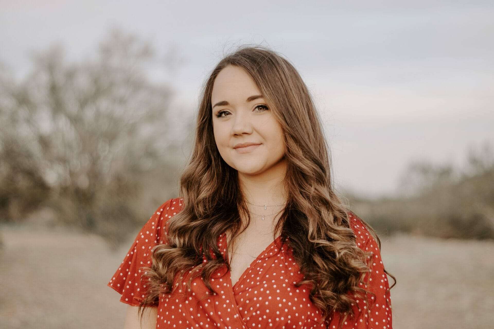 A woman with brown, wavy hair wears a red shirt with a pattern on it and smiles at the camera.