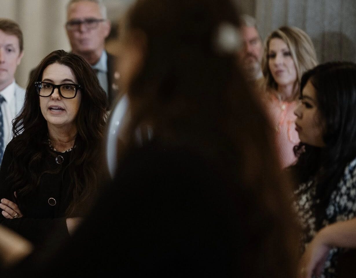 A woman with long dark hair and glasses speaks passionately to a small group in a formal indoor setting. She is surrounded by attentive listeners, including men in suits and women in business attire. The mood appears serious and engaged, suggesting a policy discussion or advocacy event.