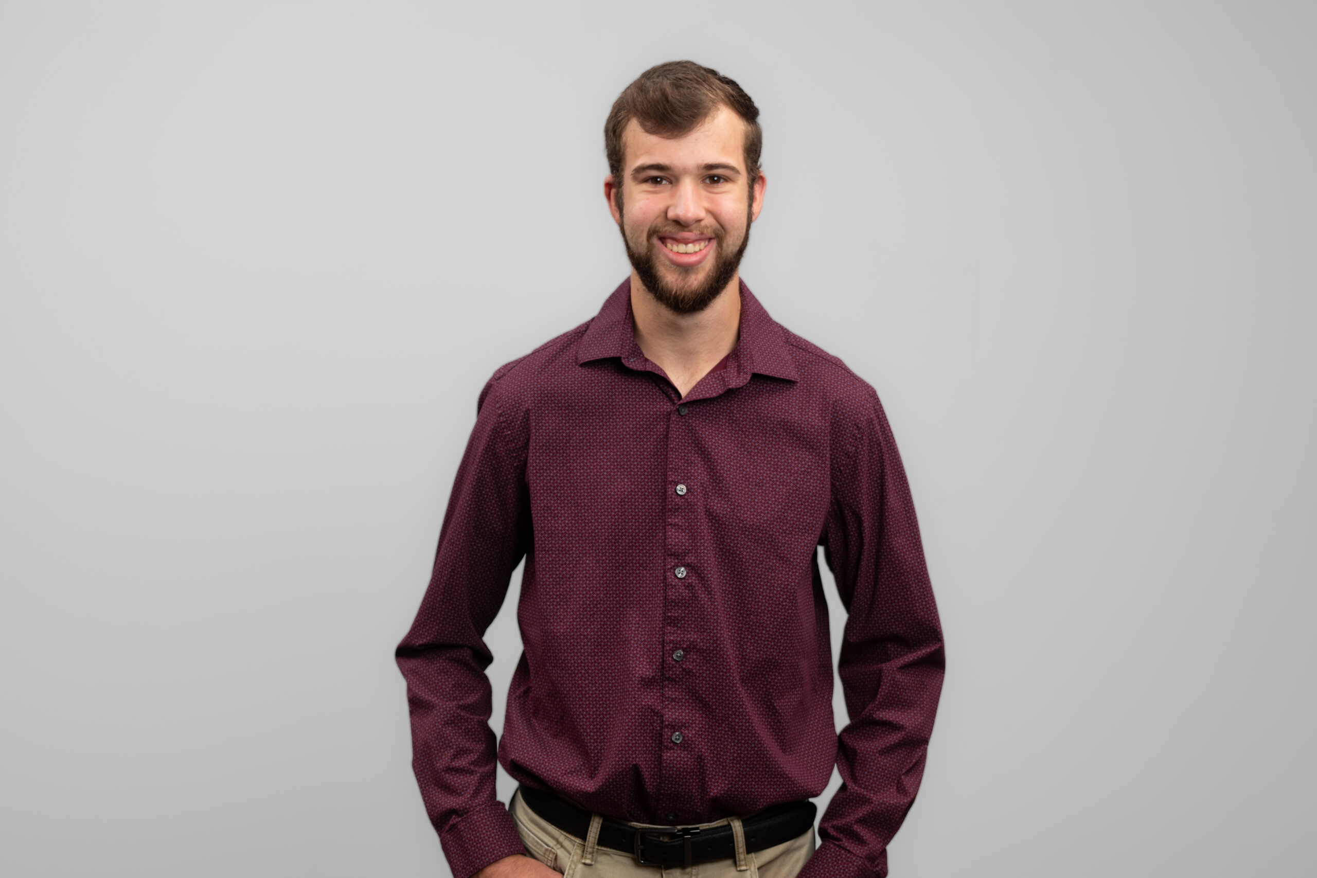 A man in a dark, collared shirt and khakis stands with his hands in his pockets and smiles at the camera.