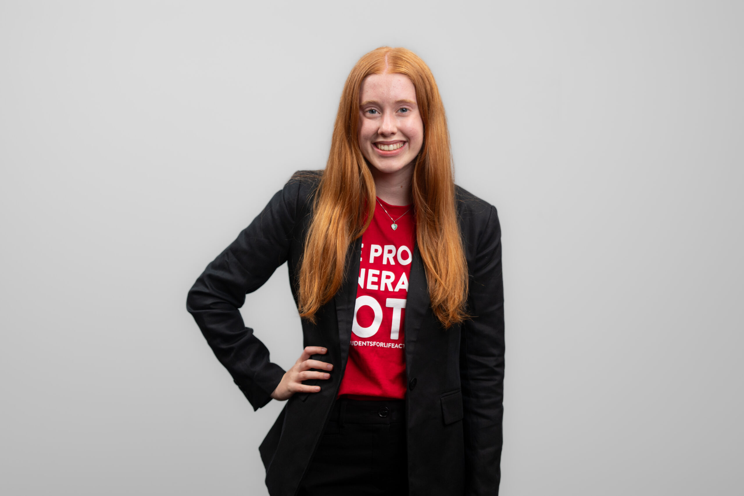A woman wears a dark suit with a t-shirt underneath and smiles at the camera while standing with her hand on her hip.
