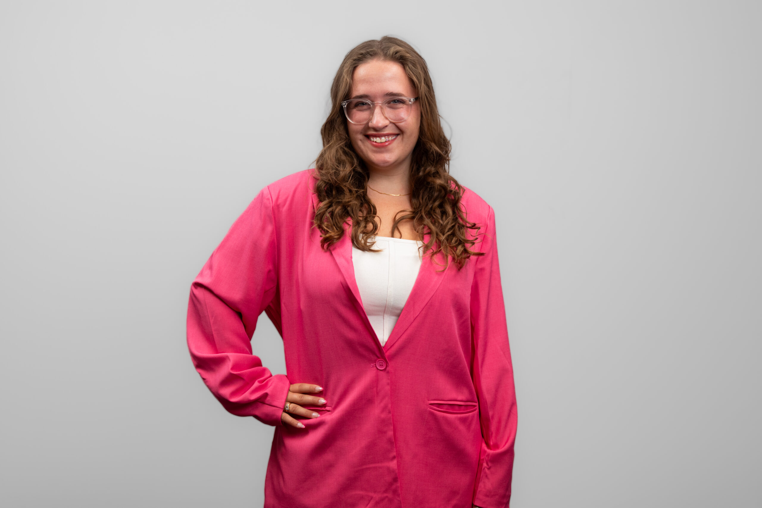 A woman in a pink outfit and white shirt underneath stands with her hand on her hip and smiles at the camera.