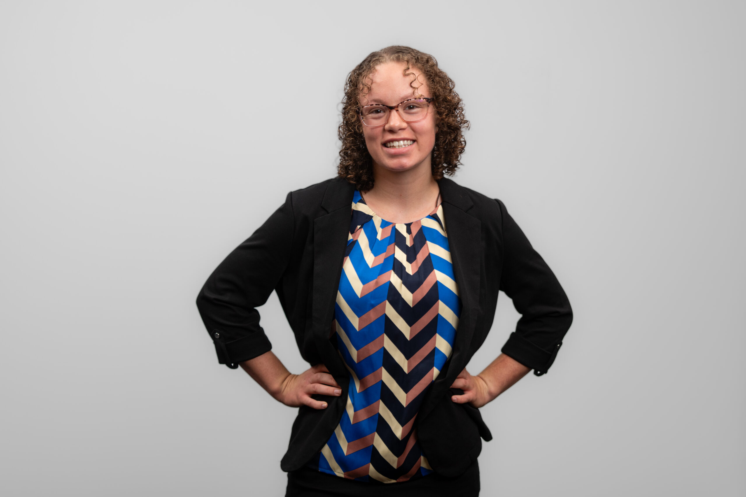 A woman with glasses stands with her hands on her hips and smiles at the camera.