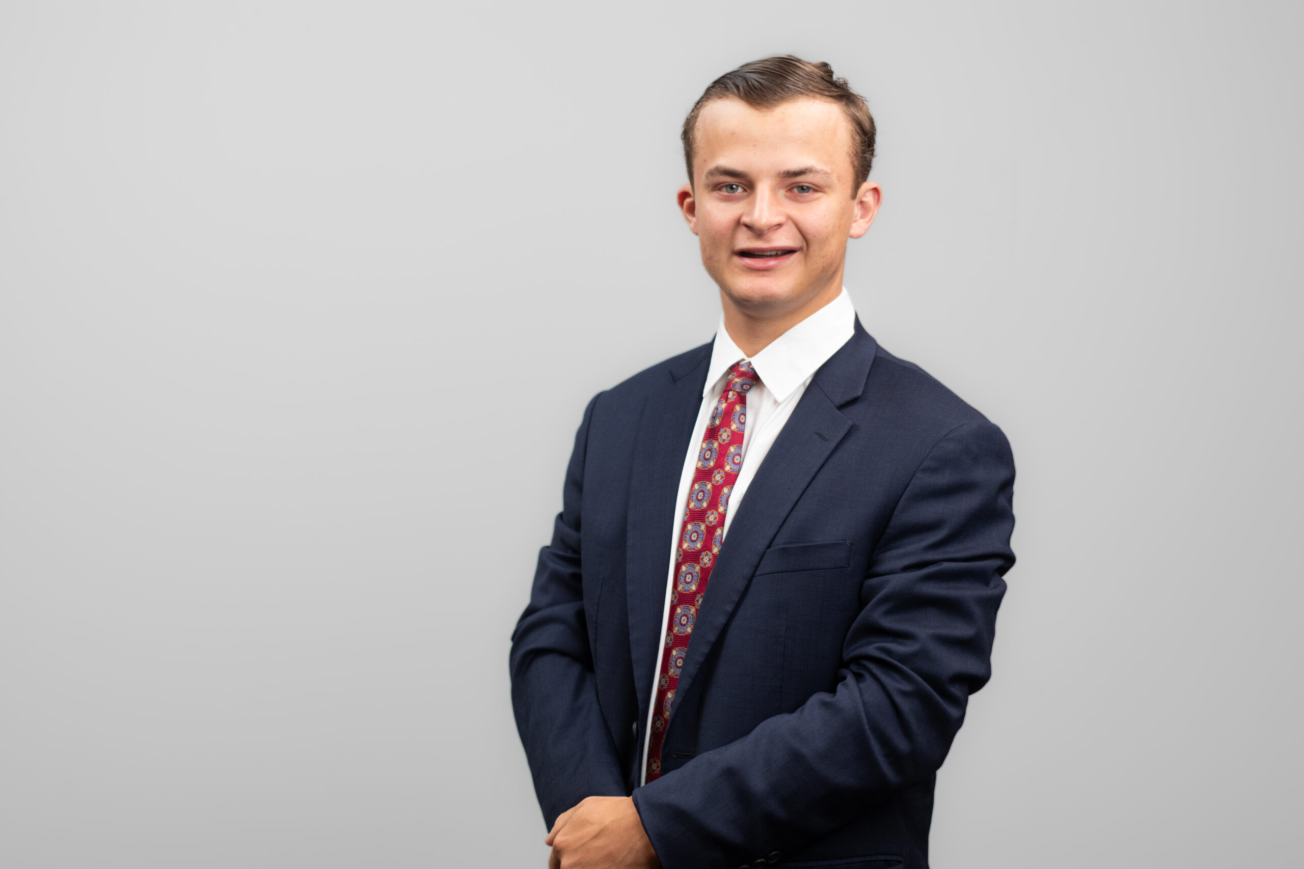 A man in a suit is turned slightly to the side. He holds one hand over the other in front of him while smiling at the camera.
