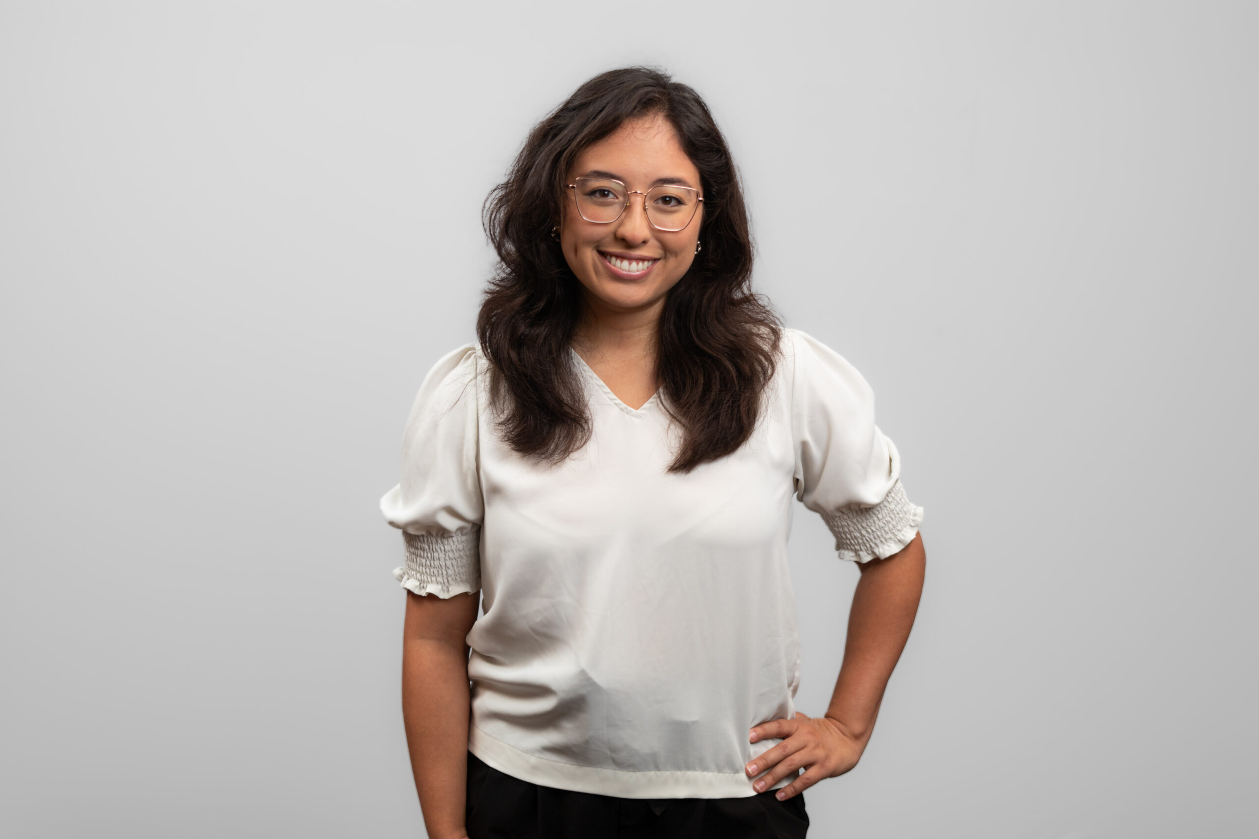 A woman wearing a white shirt stands with her hand on her hip and smiles at the camera.