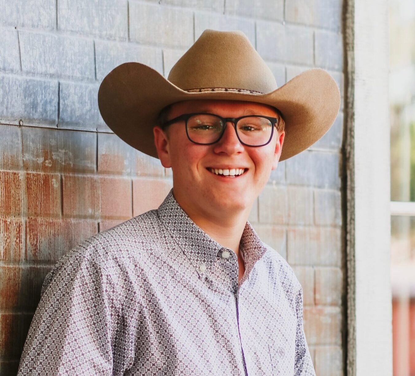 A young man in a cowboy hat, glasses, and a button-up shirt smiles at the camera.
